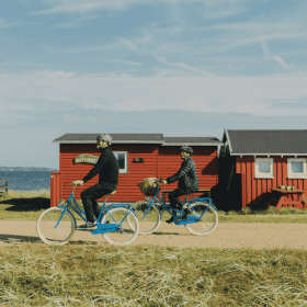 To cyklister kører på en grusvej med græs til den ene side og hav og badehuse til den anden side