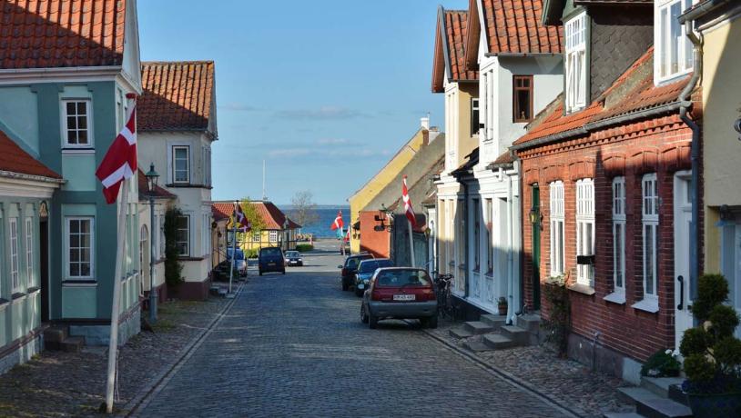 Brostensbelagt gade med gamle byhuse i forskellige farver til begge sider. Der er flag sat op foran et par af husene, et par parkerede biler, og den blå himmel ses i forsvindingspunktet.