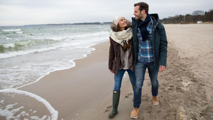 Par går langs vandkanten på stranden i tykt overtøj da det er vinter