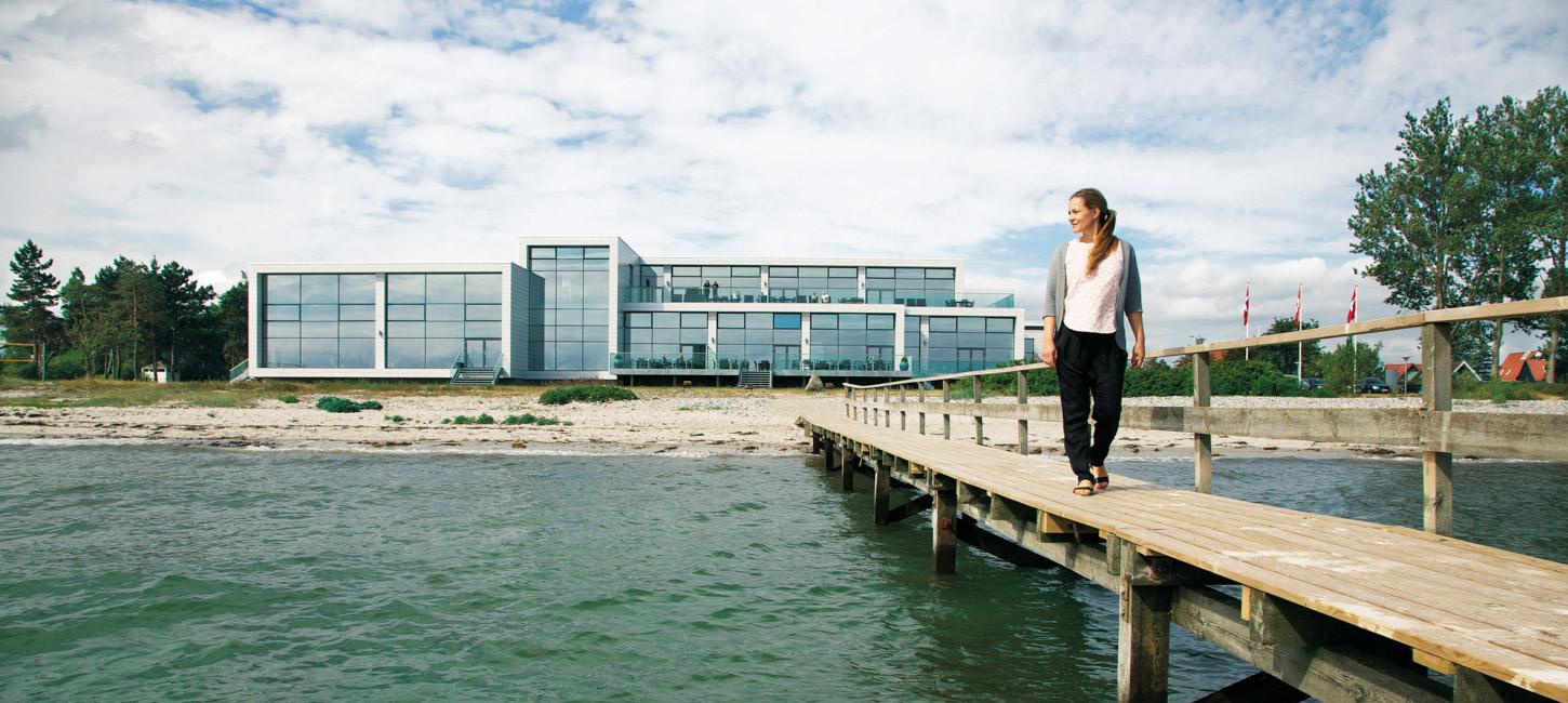 En dame går langs en badebro og kigger ud over vandet. Et stort hotel med vinduer på hele facaden ligger i baggrunden.