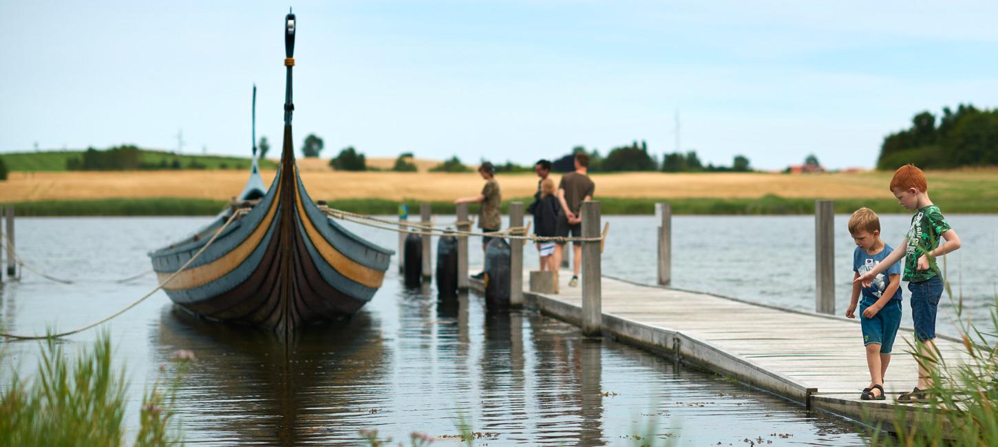 Bag lidt sløret græs spreder vandet sig, og mødes i horisonten med en mark. I højre side er en badebro. I venstre side ligger et vikingeskib. På badebroen står tre voksne og kigger på skibet, mens to børn leger i starten af badebroen.