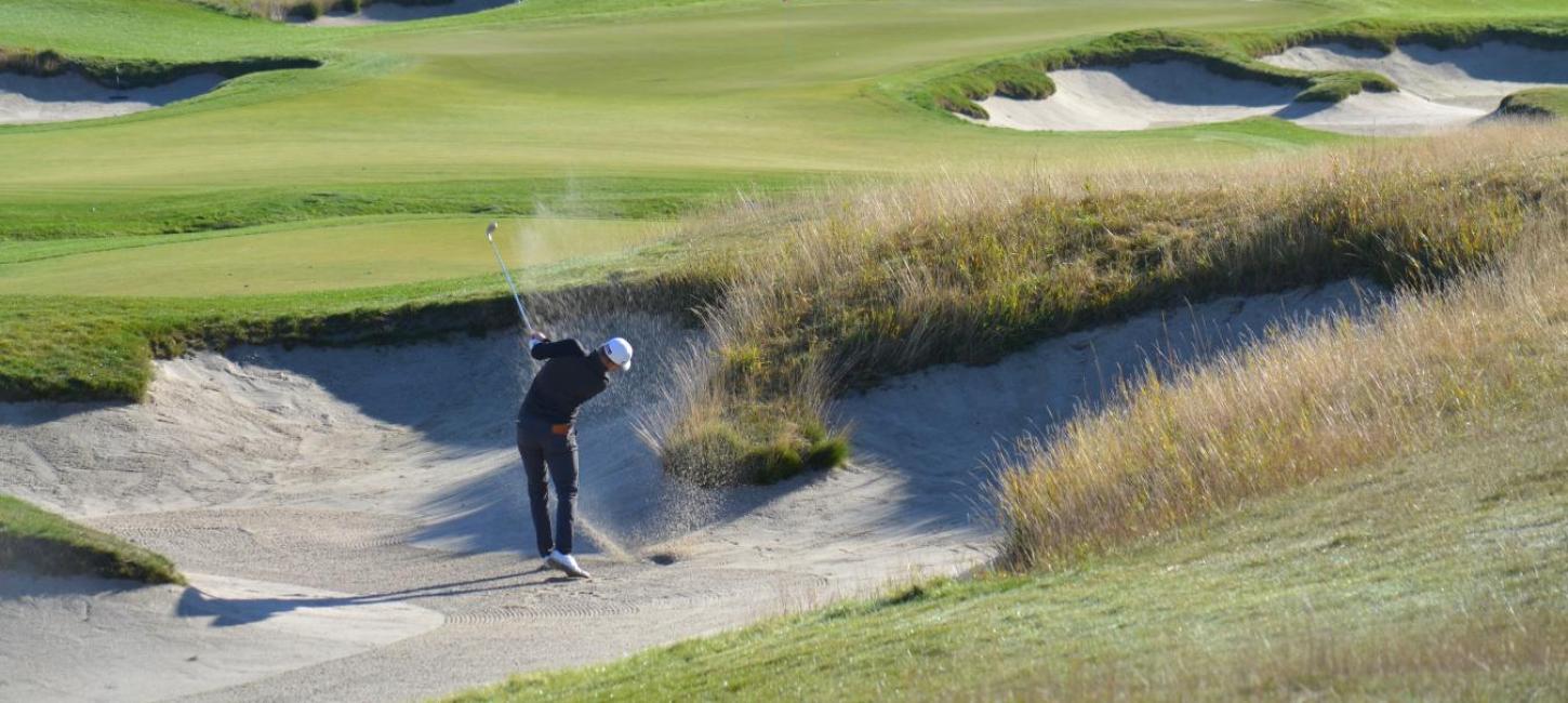 Golfspiller på bølgende grøn golfbane slår et slag fra en bunker, så sandet står op.