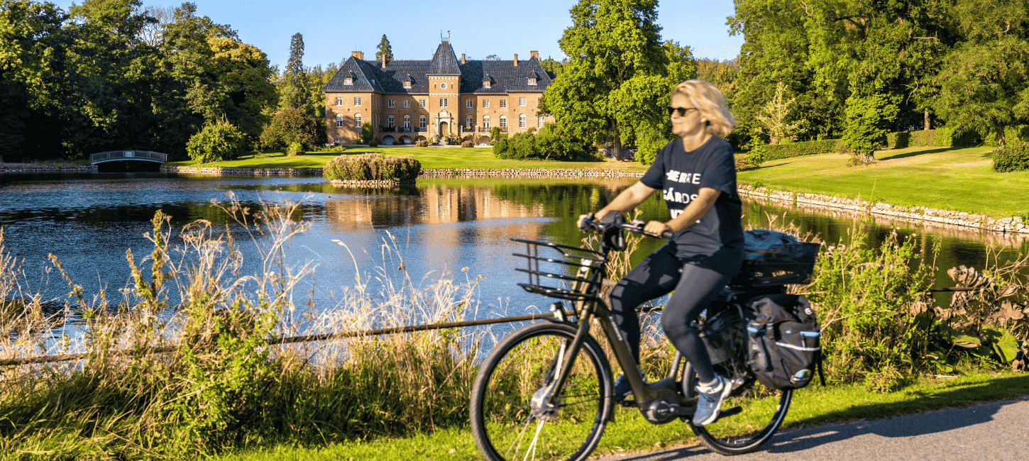 En kvinde cykler ved en sø foran et gods, omgivet af natur og sollys, nyder en fredelig dag udendørs.