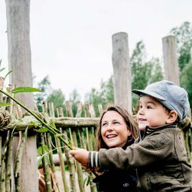 En lille dreng med grøn jakke og kasket fodrer en giraf med en bambusgren. En kvinde sidder på hug ved siden af drengen, smiler og kigger på.